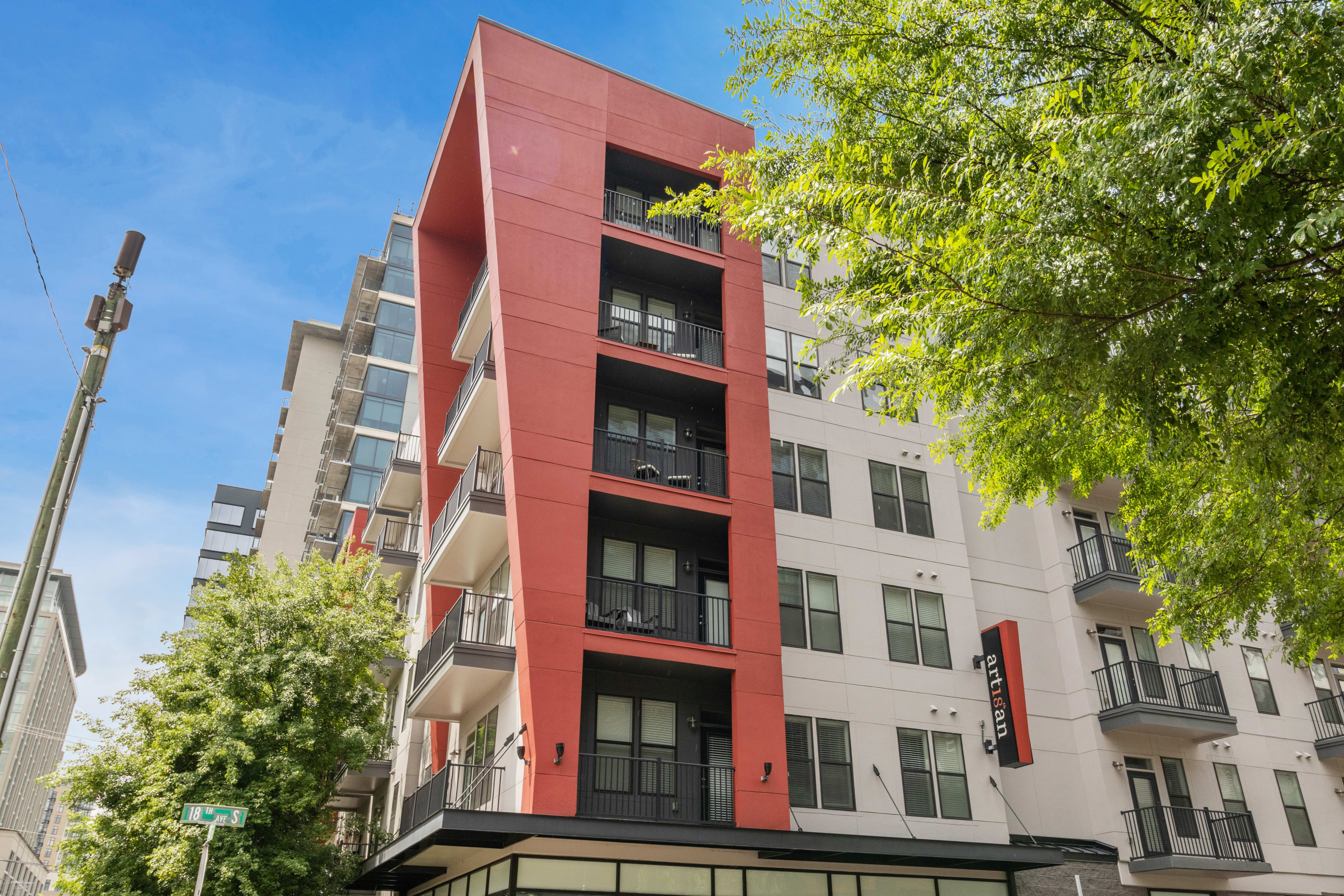 A modern multi-story apartment building with a striking red section stands behind lush green trees under a clear blue sky.