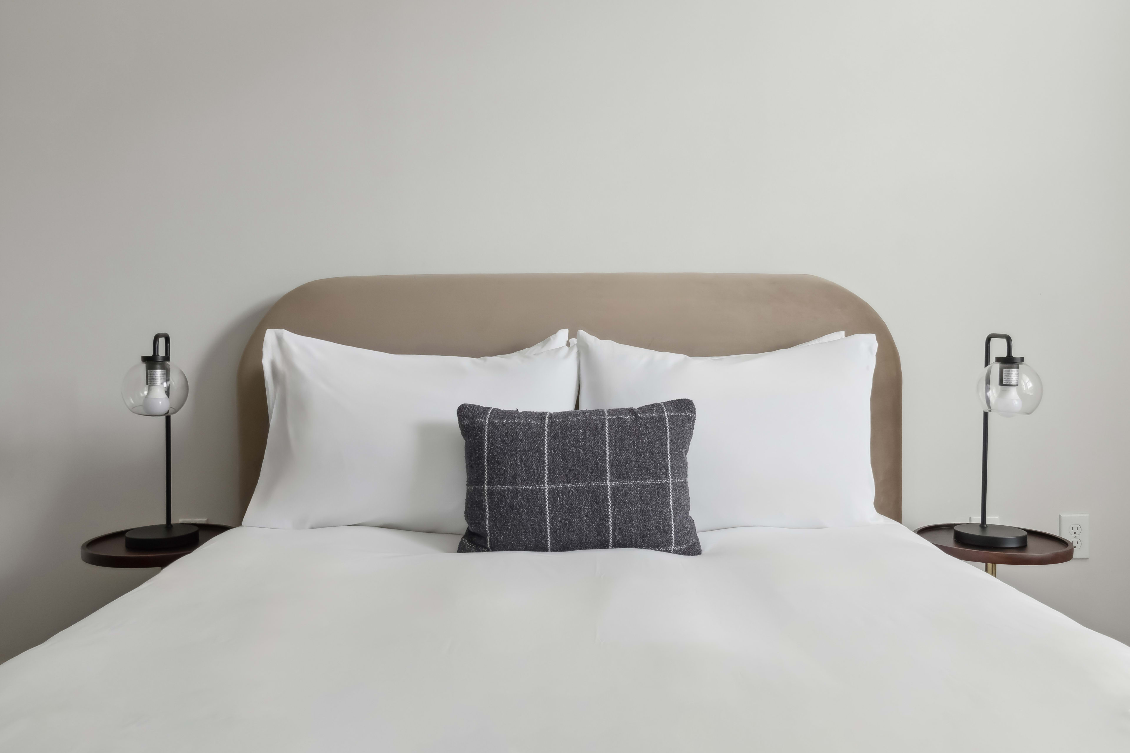 A white bed with a grey plaid pillow and two white pillows sits against a beige headboard, flanked by two small side tables with modern lamps against a plain white wall.