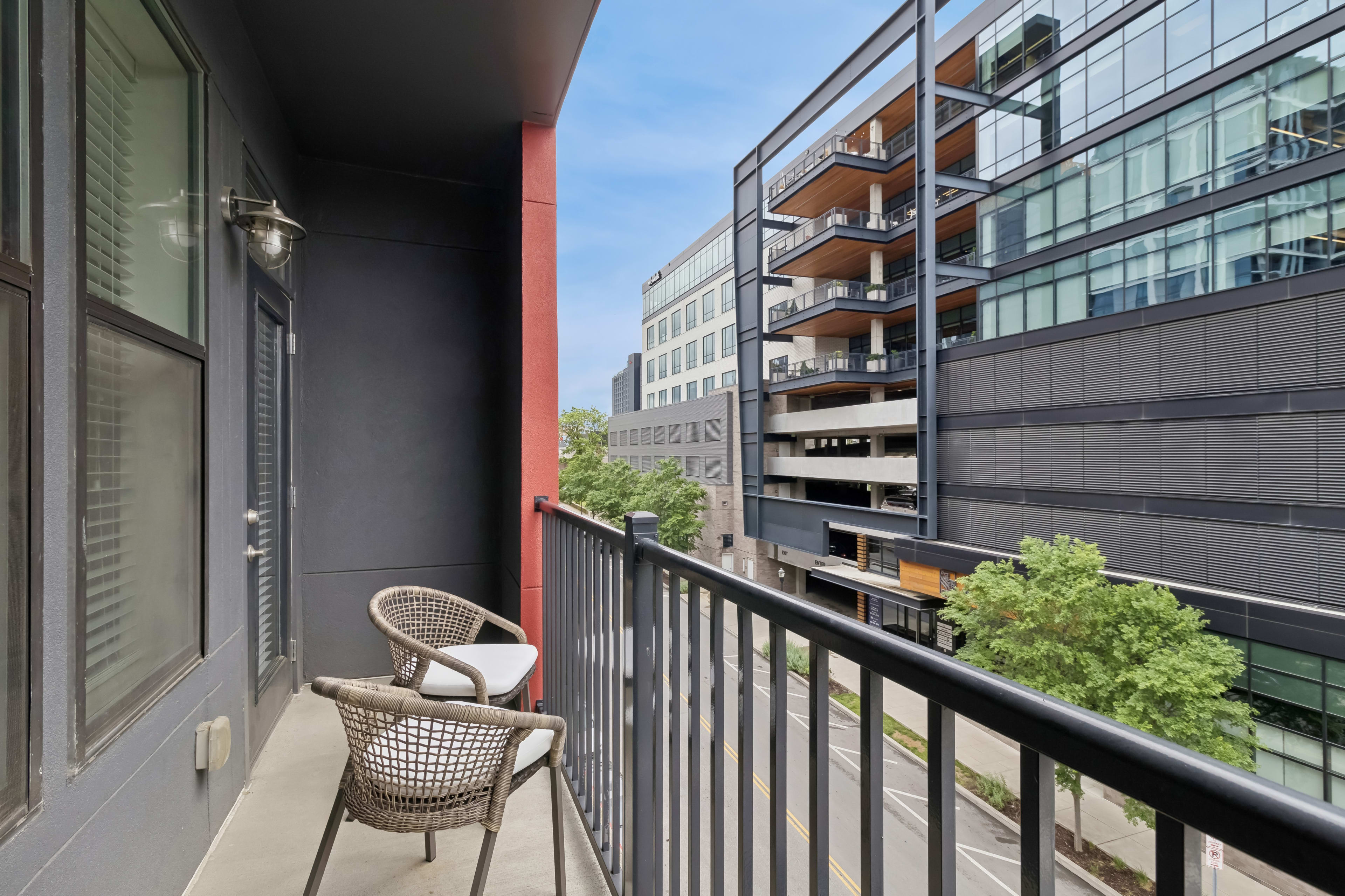 Two wicker chairs sit on a grey balcony in the foreground, overlooking a street and modern glass buildings in the background.