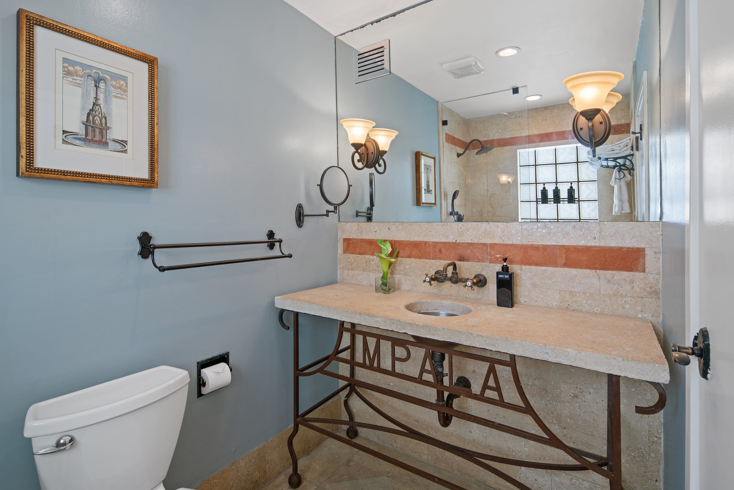A white toilet and a stone sink with a decorative metal base sit in the foreground of a blue-walled bathroom featuring a large mirror and wall sconces in the background.