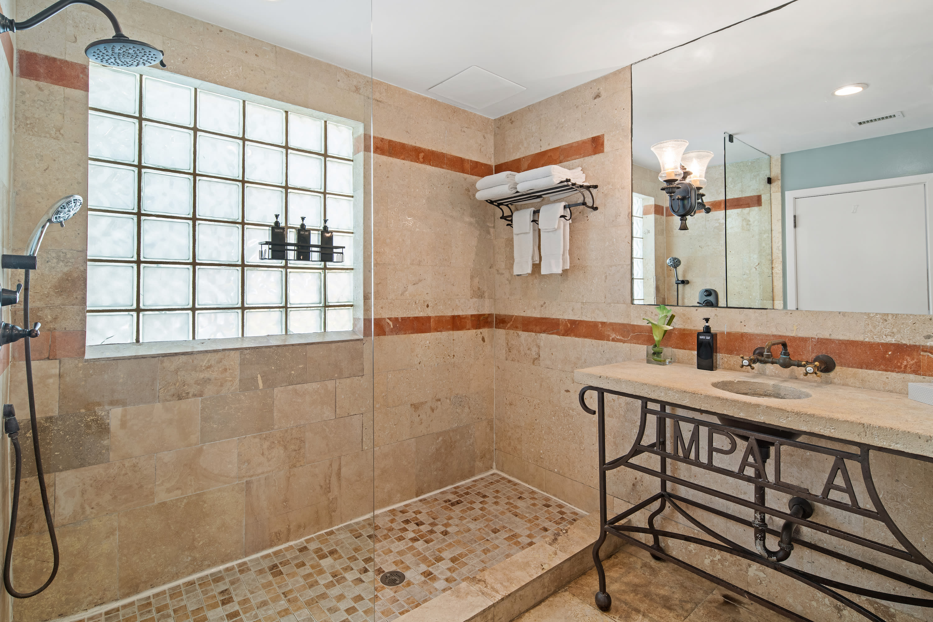 A walk-in shower with a glass partition and glass block window stands in the foreground, while a stone vanity with a decorative metal base and a large mirror are in the background.