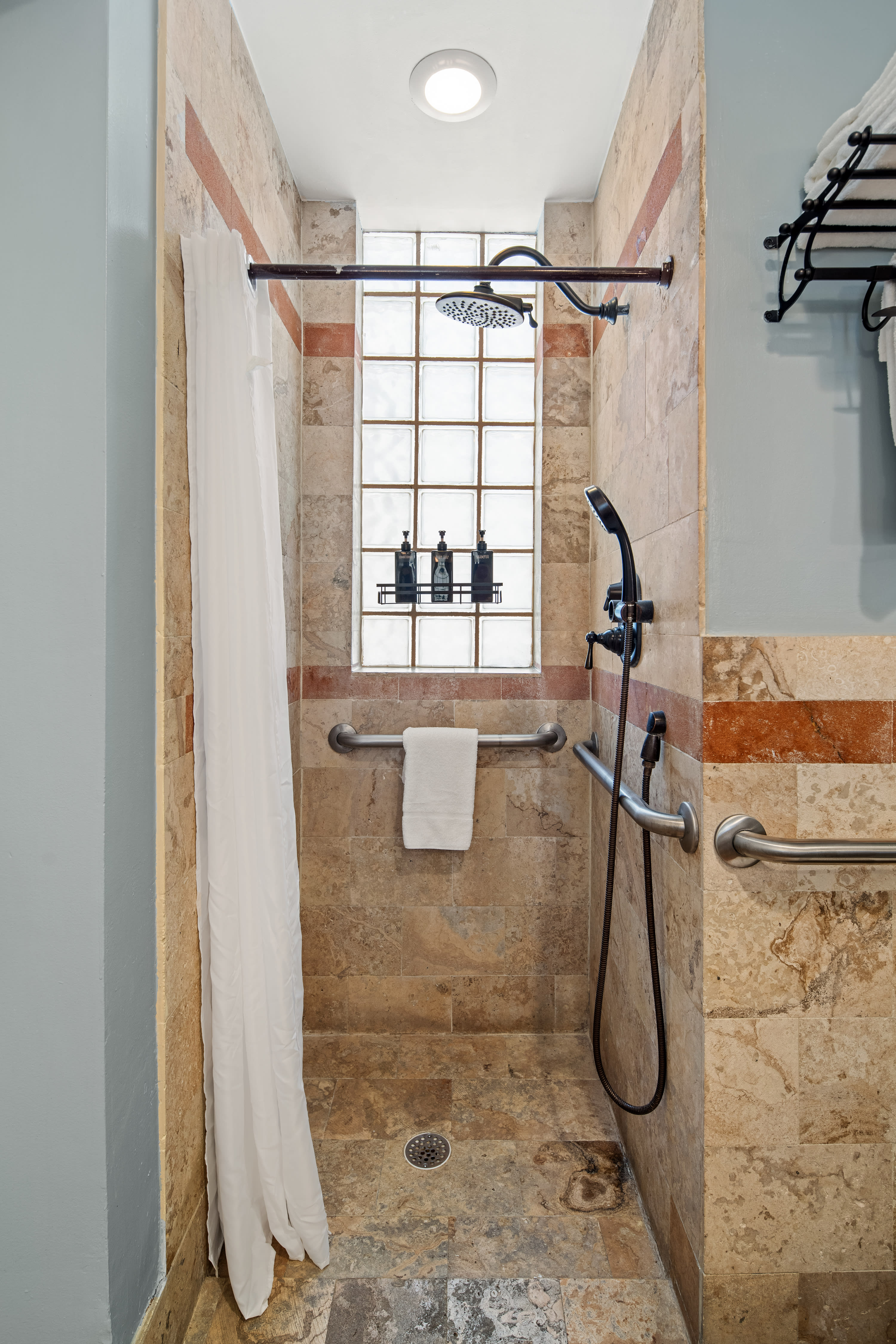 A walk-in shower with tan stone tiles and grab bars features a white curtain in the foreground and a glass block window in the background.