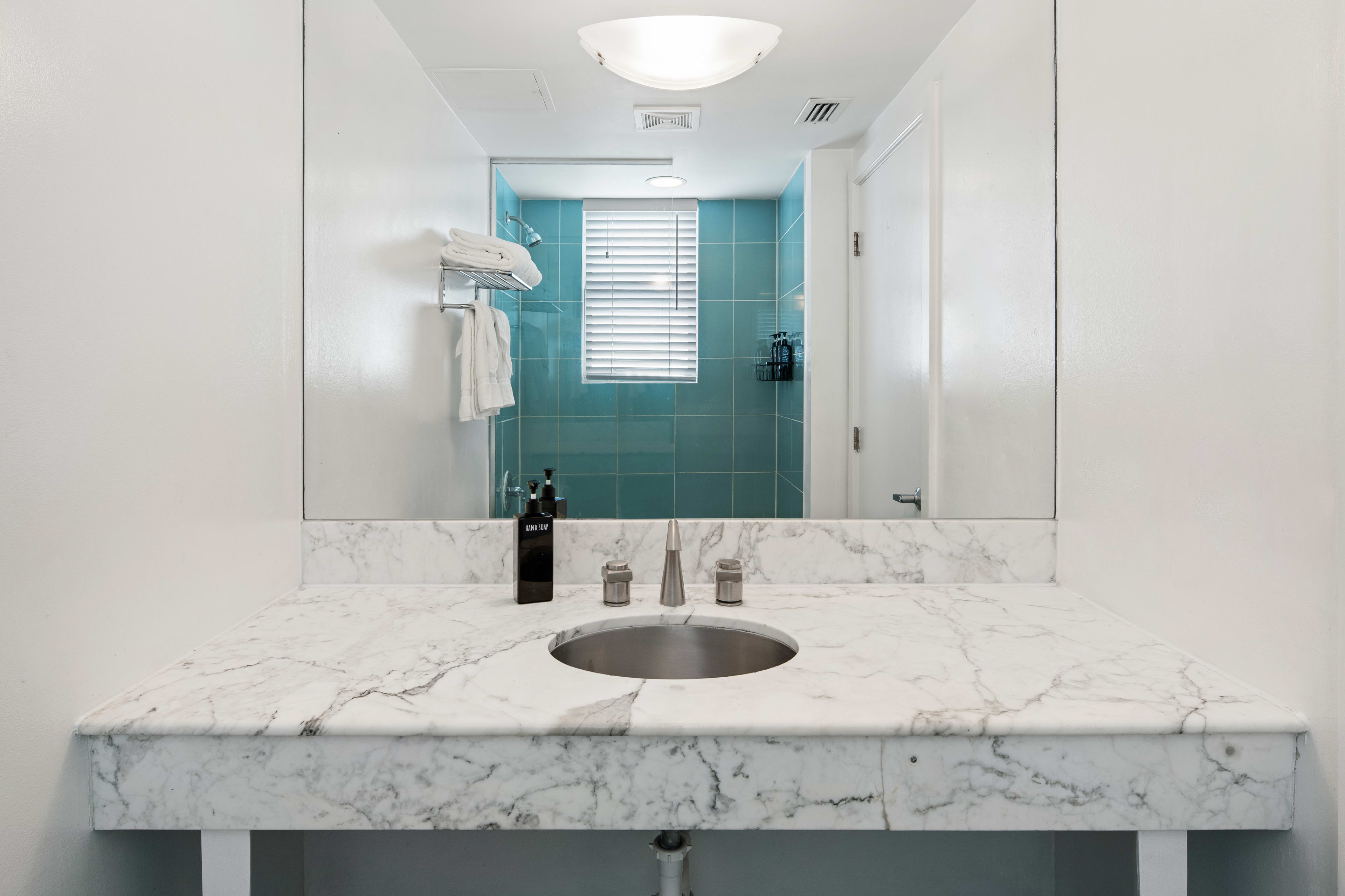 A white marble countertop with a built-in sink sits in the foreground, while a large mirror reflects a bathroom with blue tiles and a window in the background.