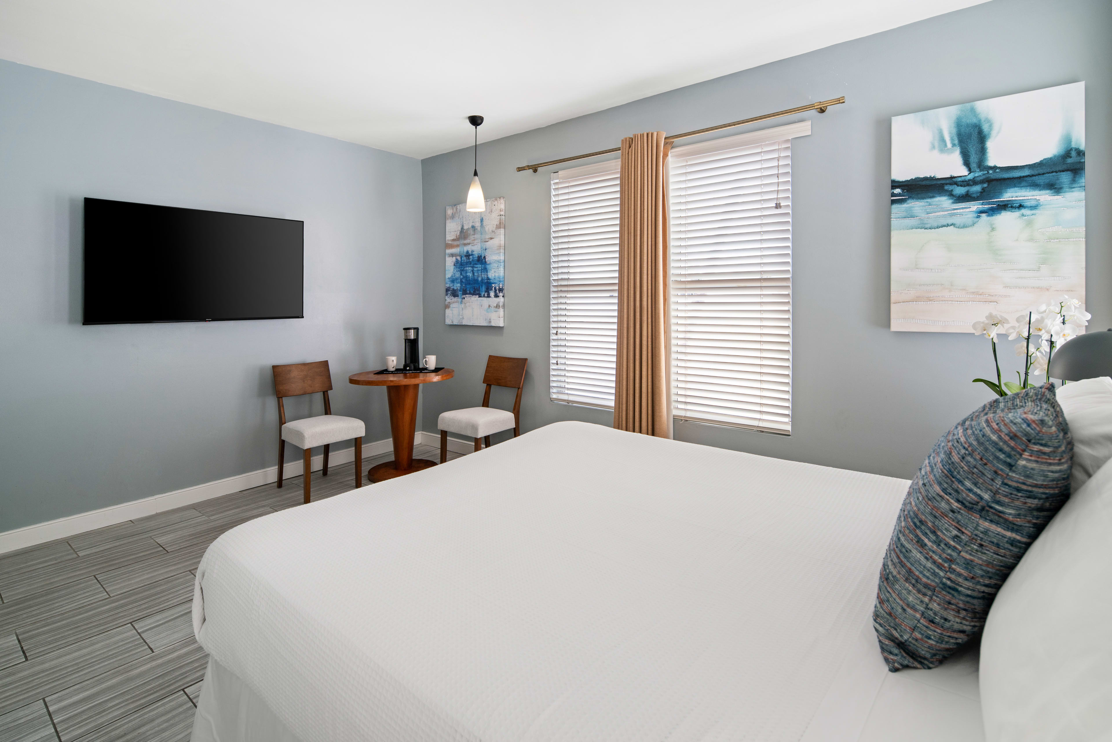 A white bed occupies the foreground of a modern bedroom featuring a small dining set, a wall-mounted TV, and a window with beige curtains in the background.