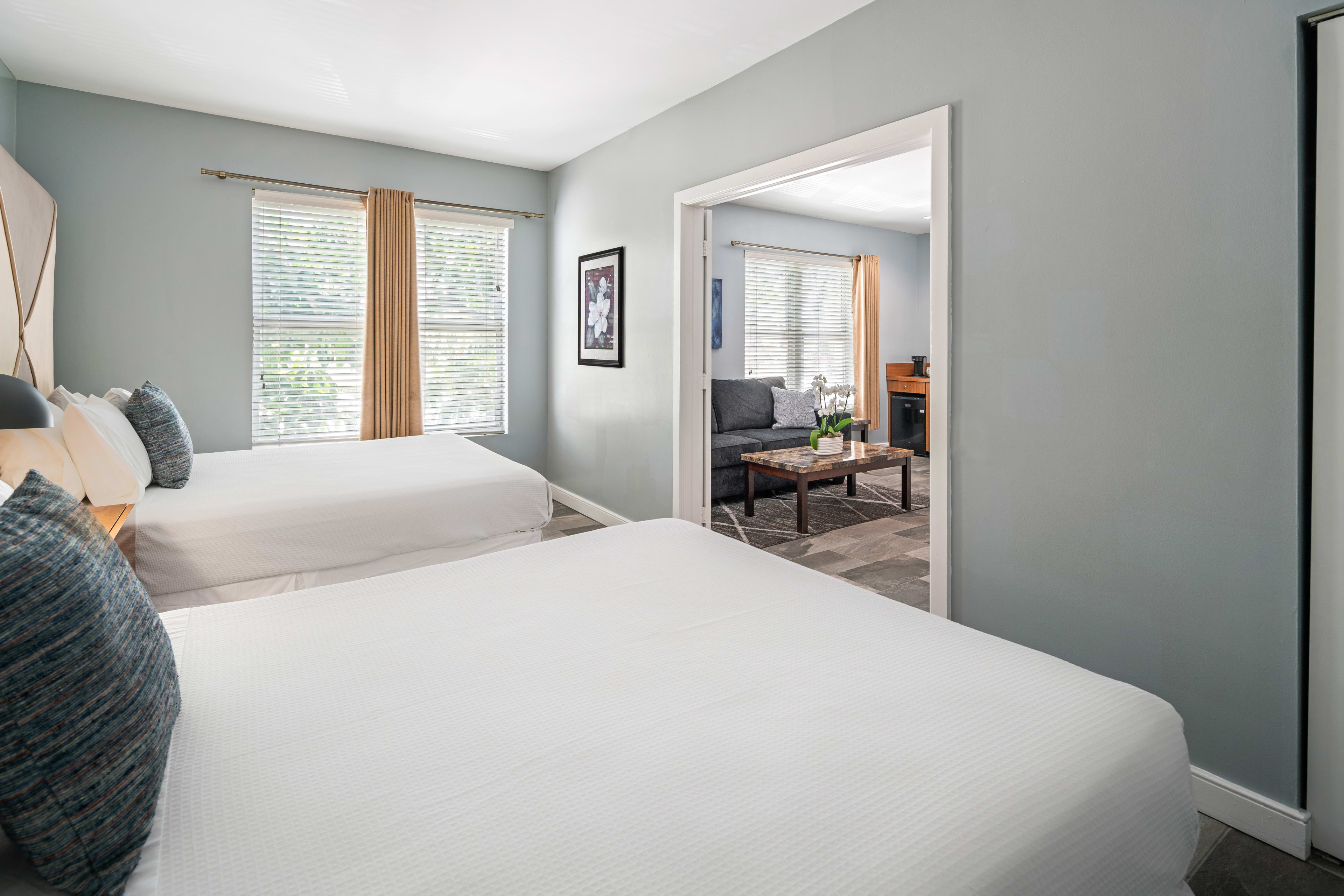 Two white beds with blue pillows occupy the foreground of a light blue room, with a doorway leading to a living area featuring a grey sofa and coffee table in the background.
