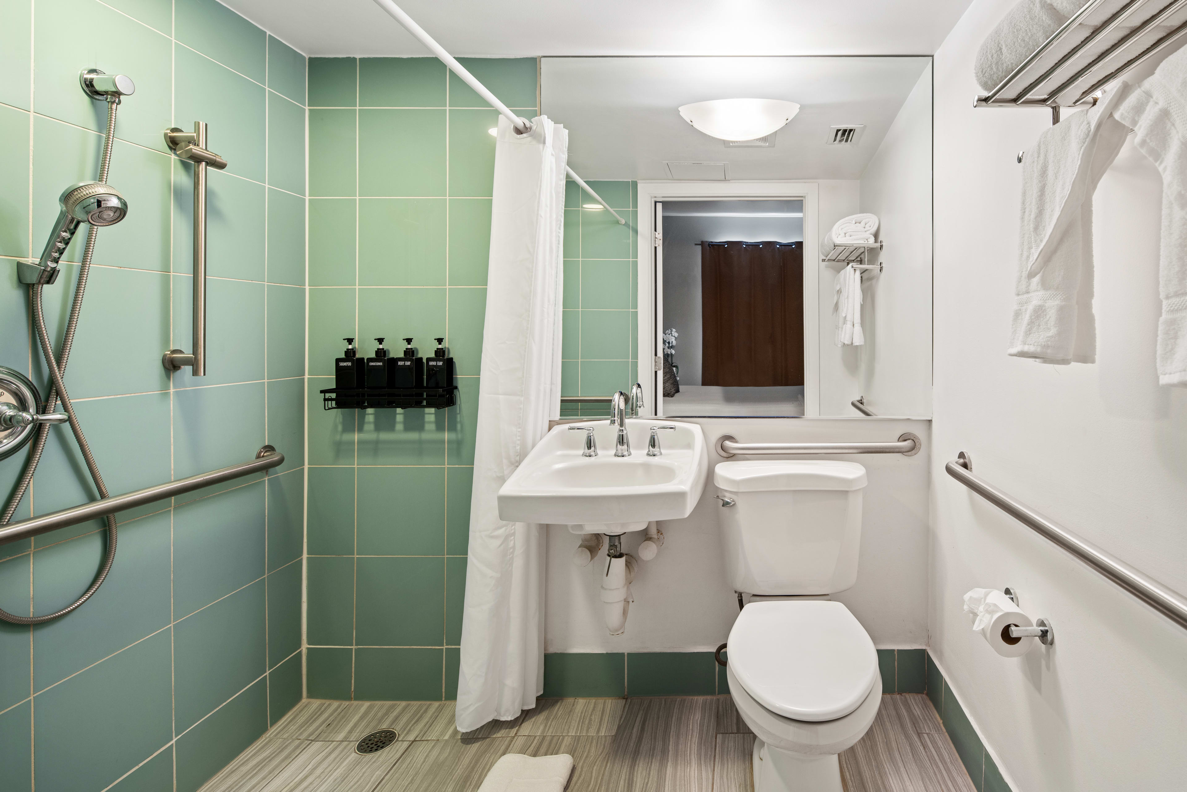 A white toilet and sink with safety grab bars sit in the foreground of a bathroom featuring a green-tiled shower area and a large mirror in the background.