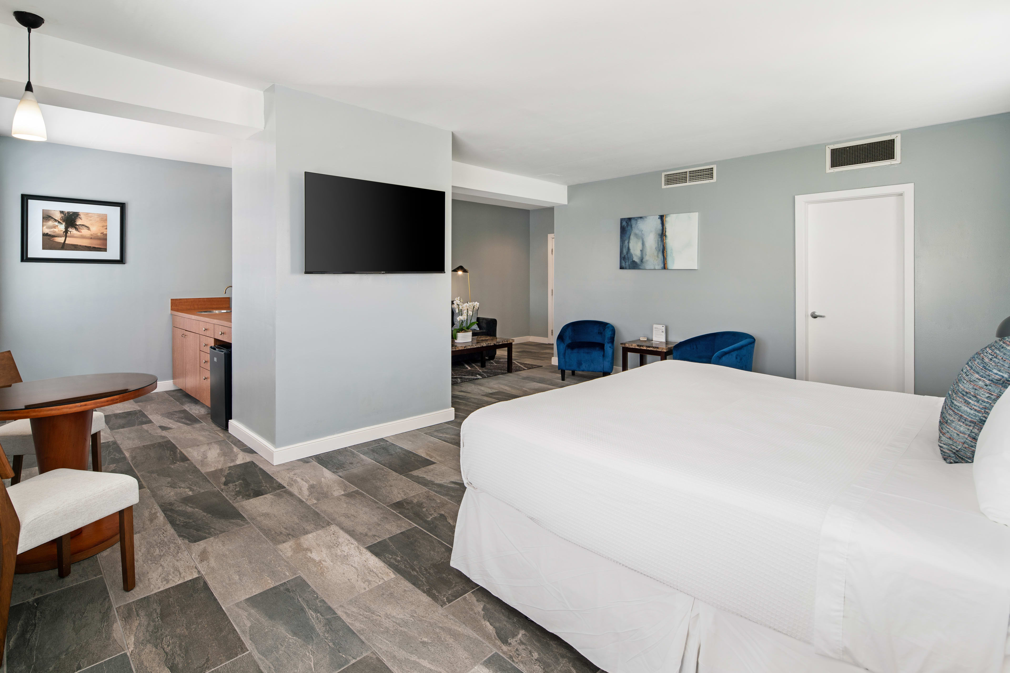 A white bed occupies the foreground of a modern hotel room featuring a wall-mounted television and a seating area with blue chairs in the background.
