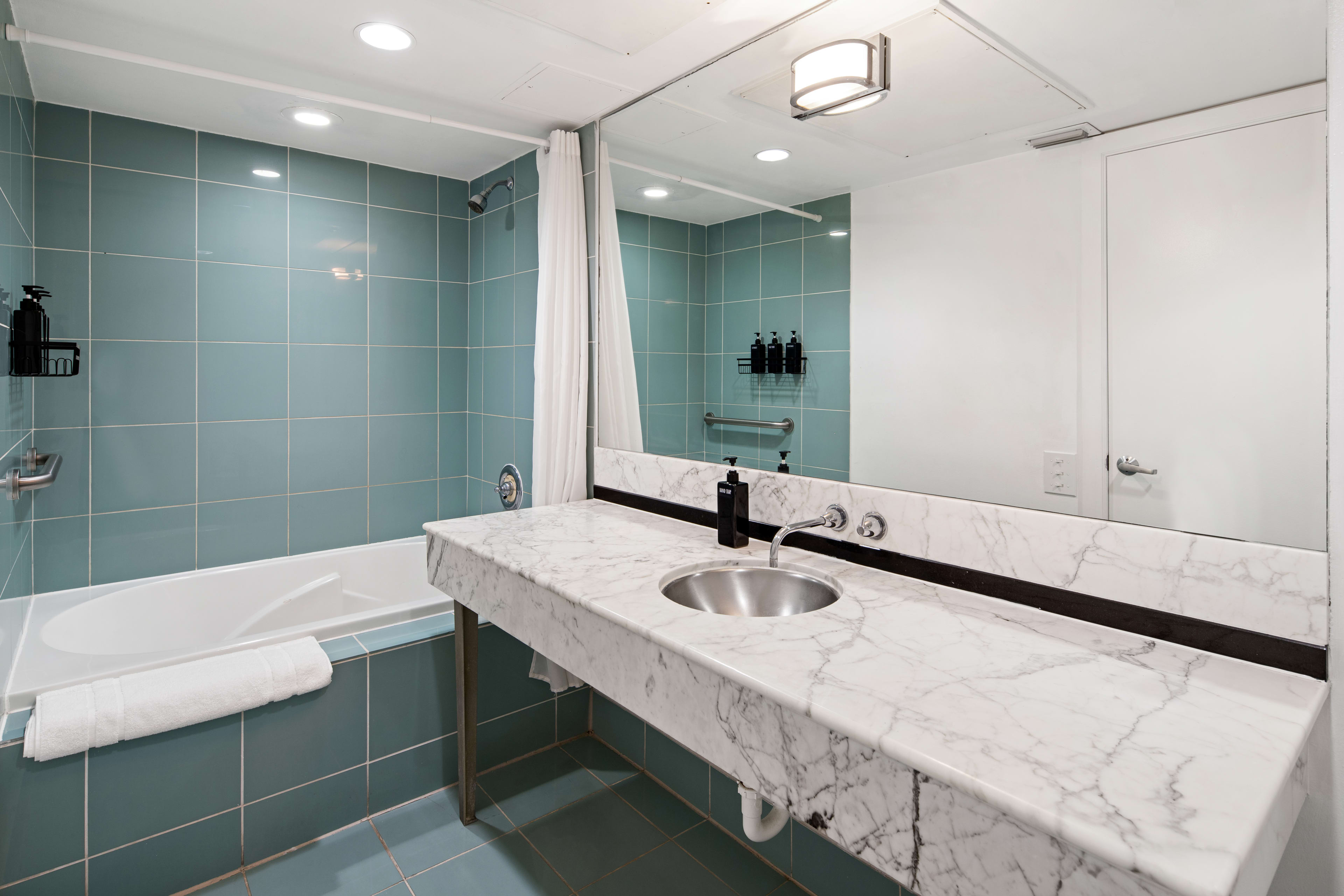 A white marble countertop with a sink sits in the foreground, while a blue-tiled bathtub and a large mirror occupy the background.
