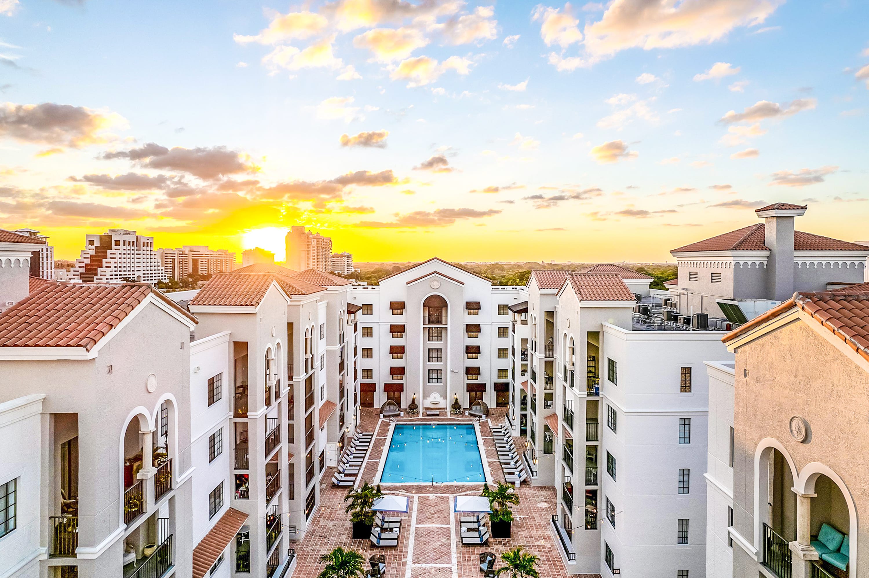 White multi-story apartment buildings surround a central courtyard with a swimming pool, set against a city skyline under a glowing sunset sky.