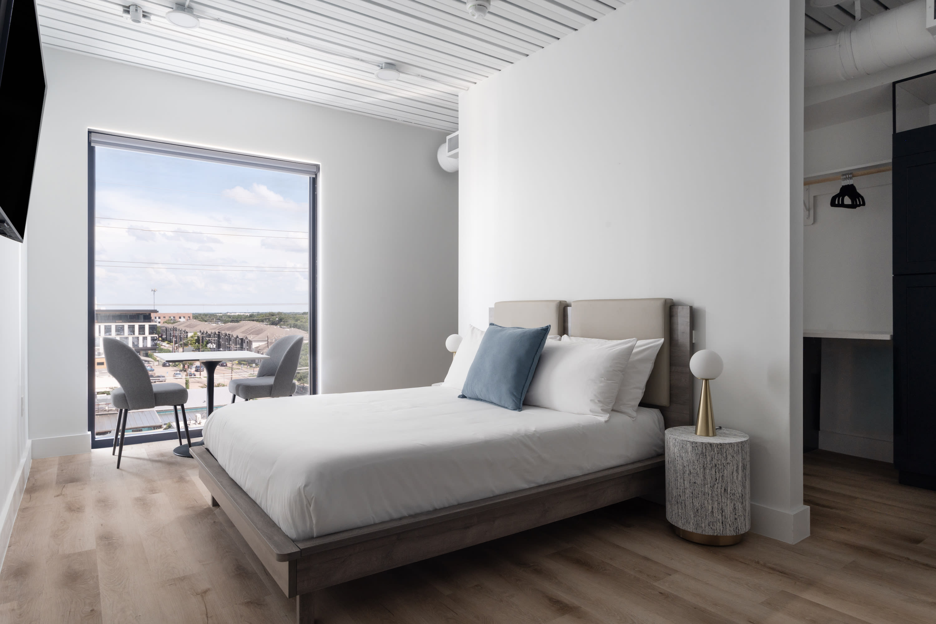 A neatly made bed with blue and white pillows sits in a modern white room, with a small dining set positioned before a large window overlooking a residential area.
