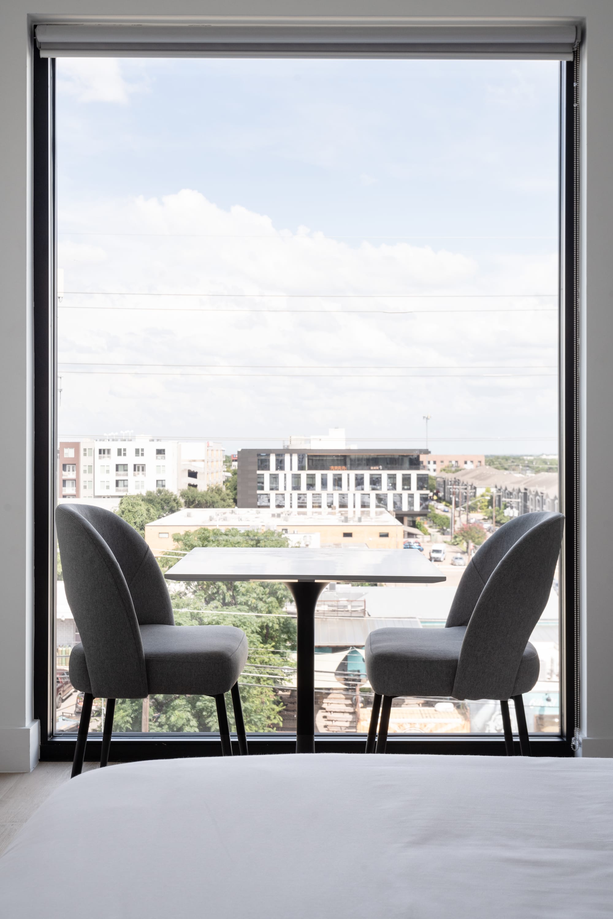 Two grey chairs and a small white table sit before a large window overlooking a city skyline under a cloudy sky.