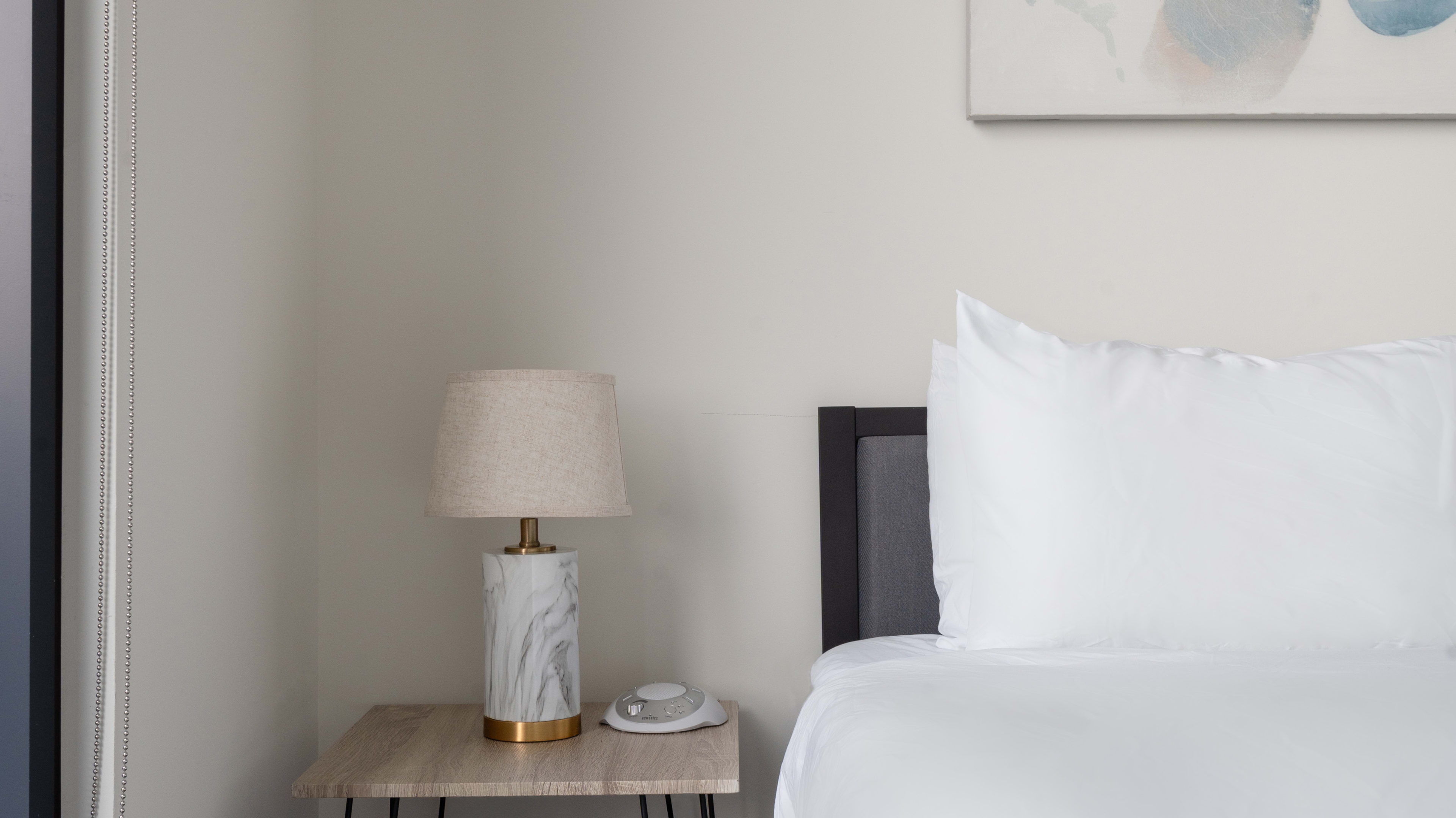A marble-based lamp and a small white clock sit on a wooden nightstand next to a bed with white linens against a plain wall.