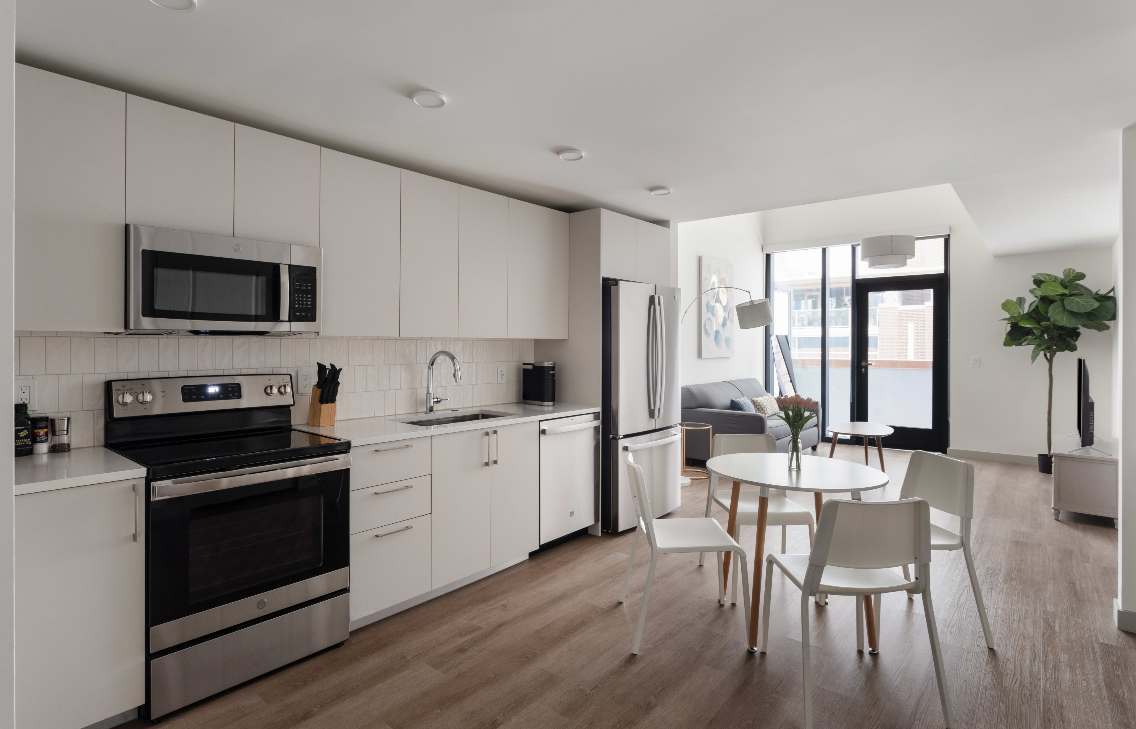 A modern white kitchen with stainless steel appliances occupies the foreground, leading into a bright living area with a small dining table and a grey sofa in the background.