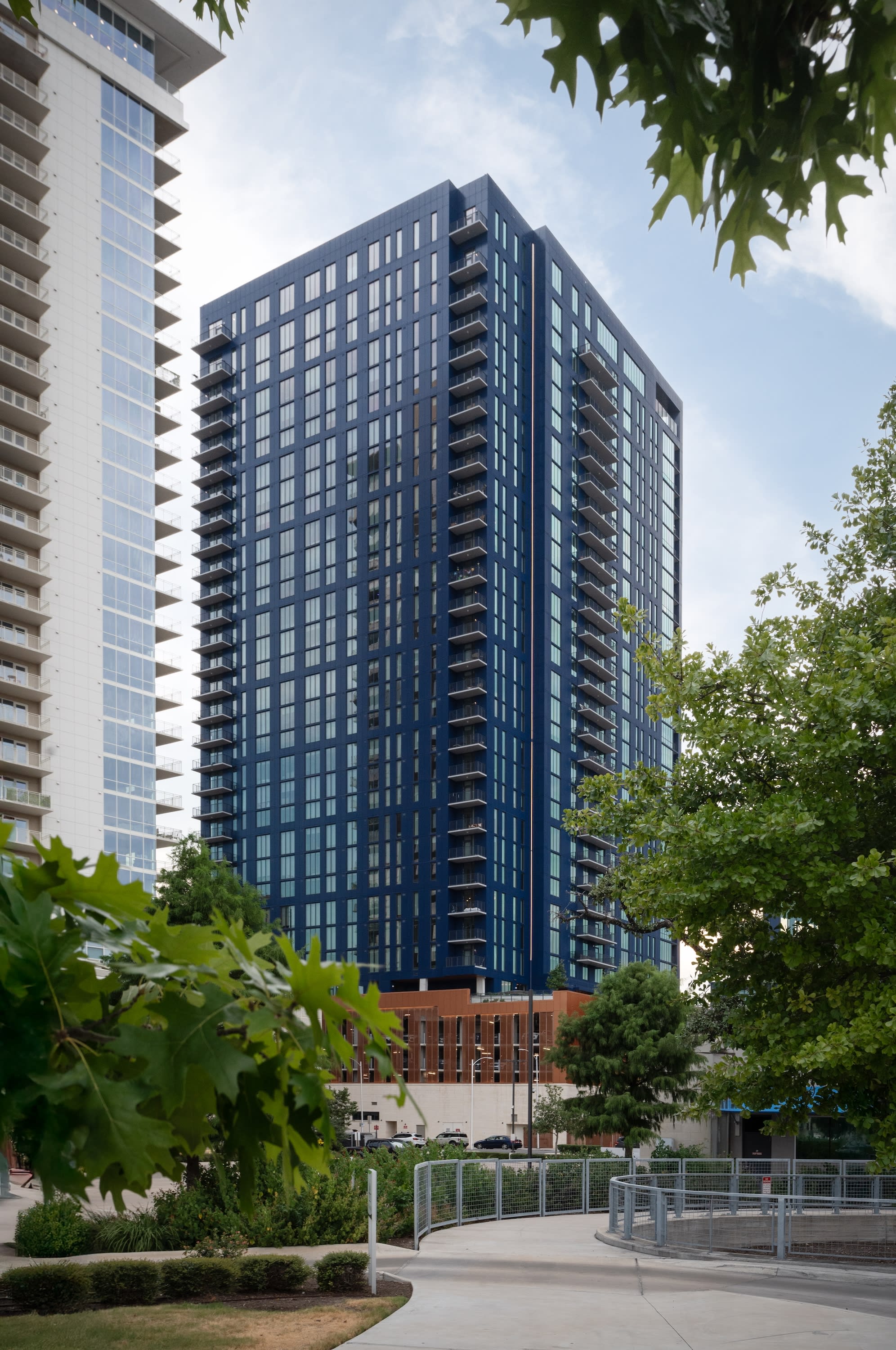 Lush green foliage frames a view of a modern blue glass skyscraper and a white high-rise building under a clear sky.