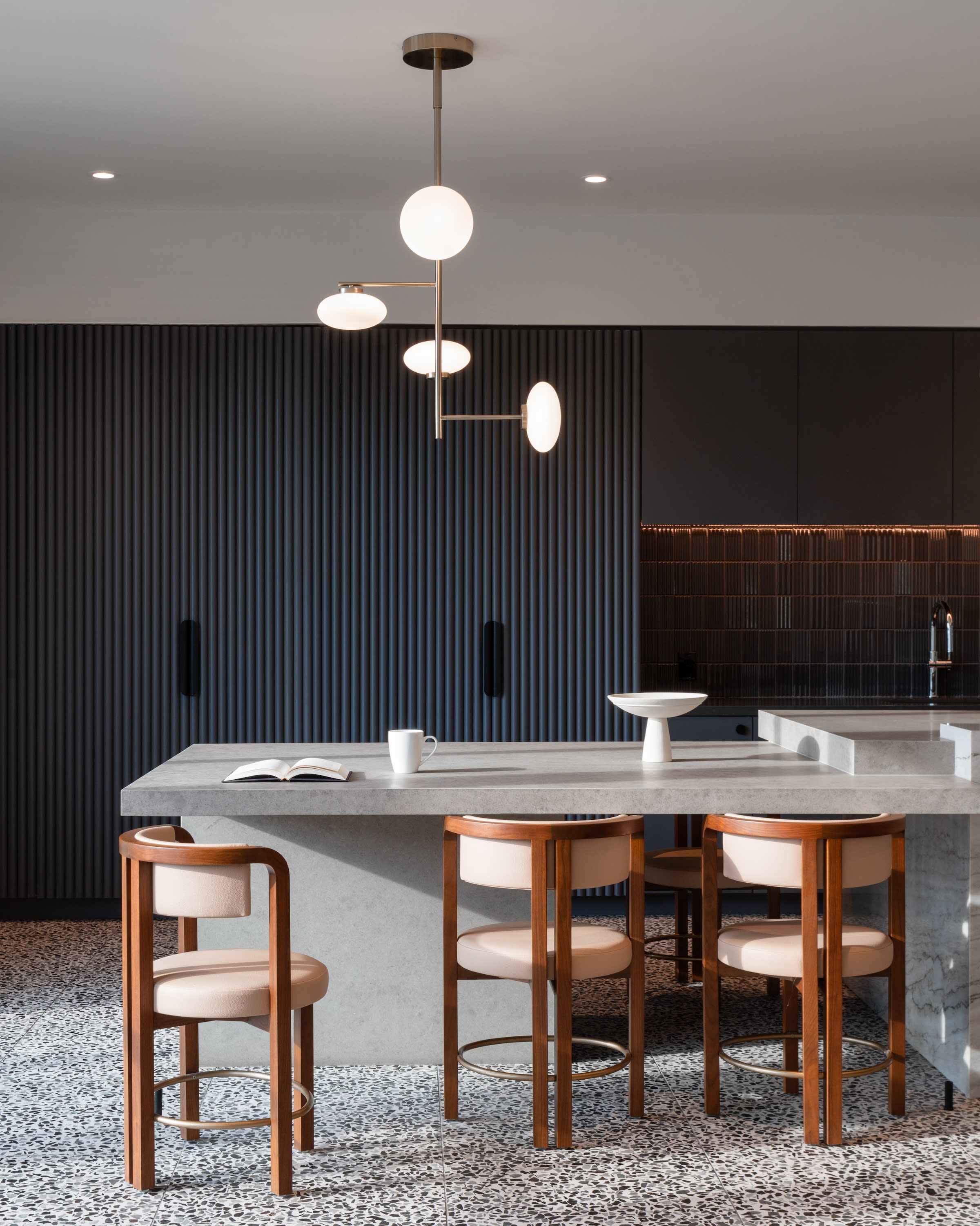 Three modern wooden bar stools sit before a grey concrete kitchen island, set against a backdrop of dark slatted walls and a minimalist black kitchen.