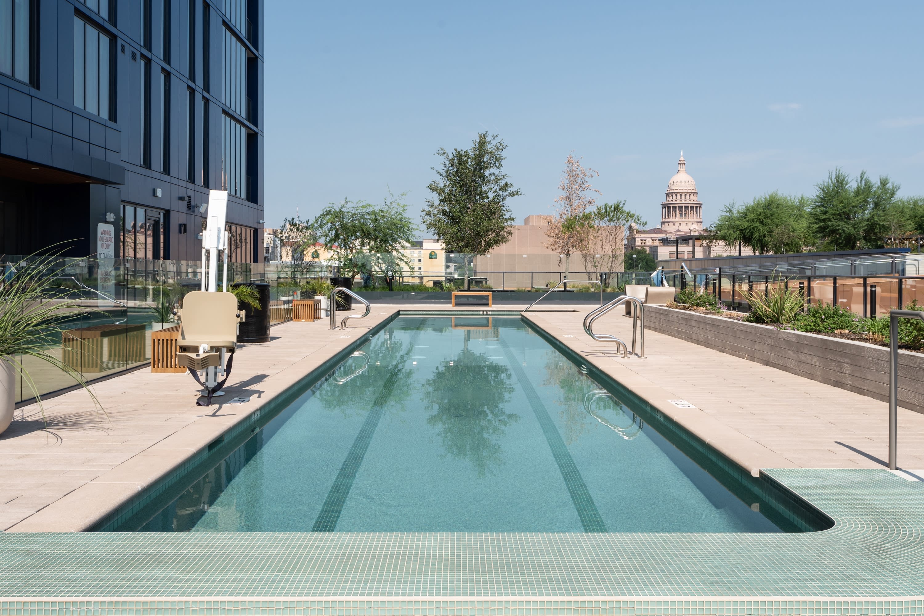 A long lap pool sits on a rooftop terrace next to a modern building, with a city skyline and a domed capitol building in the distance.