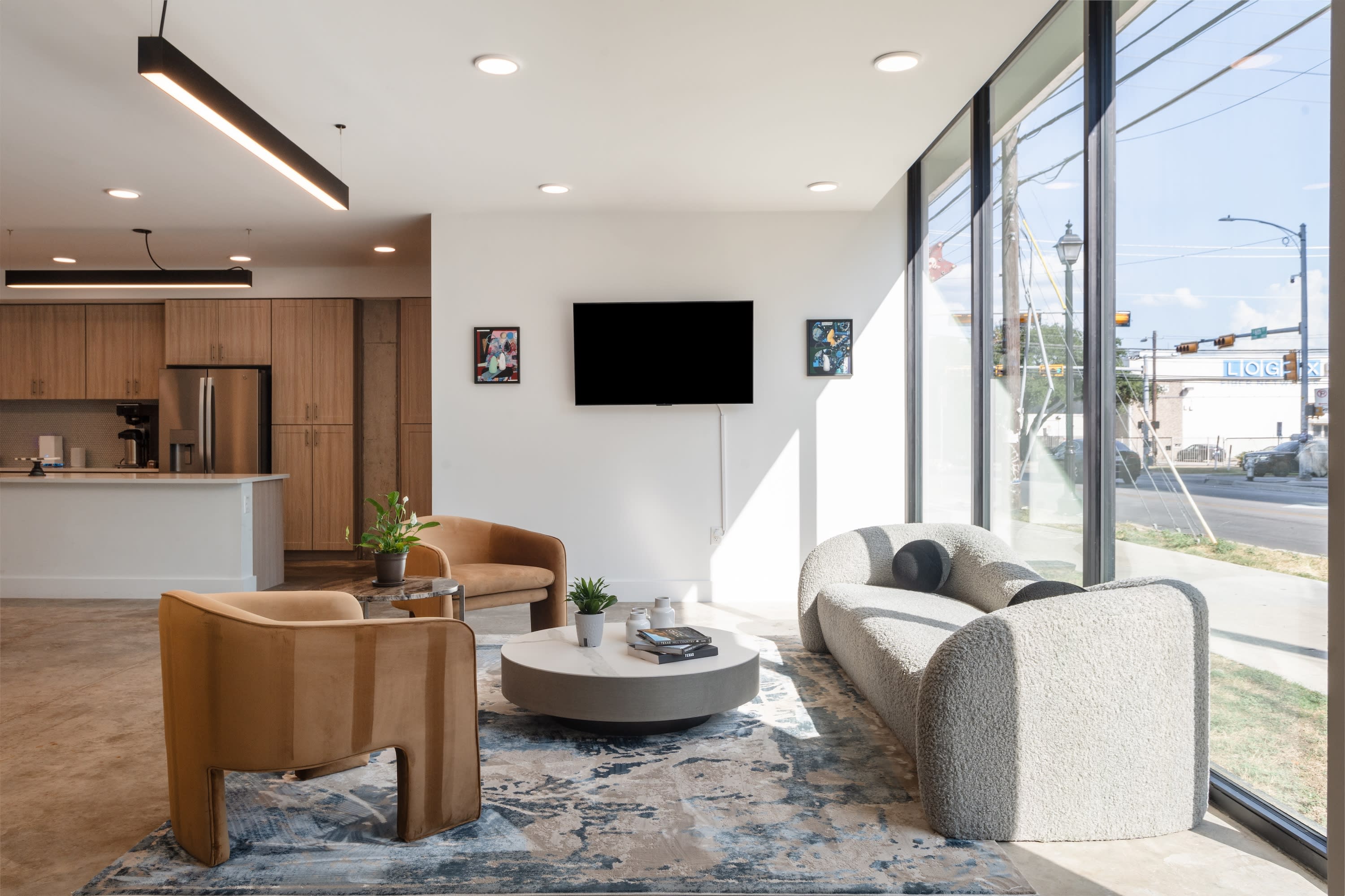 A modern living area with a curved white sofa and tan armchairs sits in the foreground, while a kitchen and large windows overlooking a street are in the background.