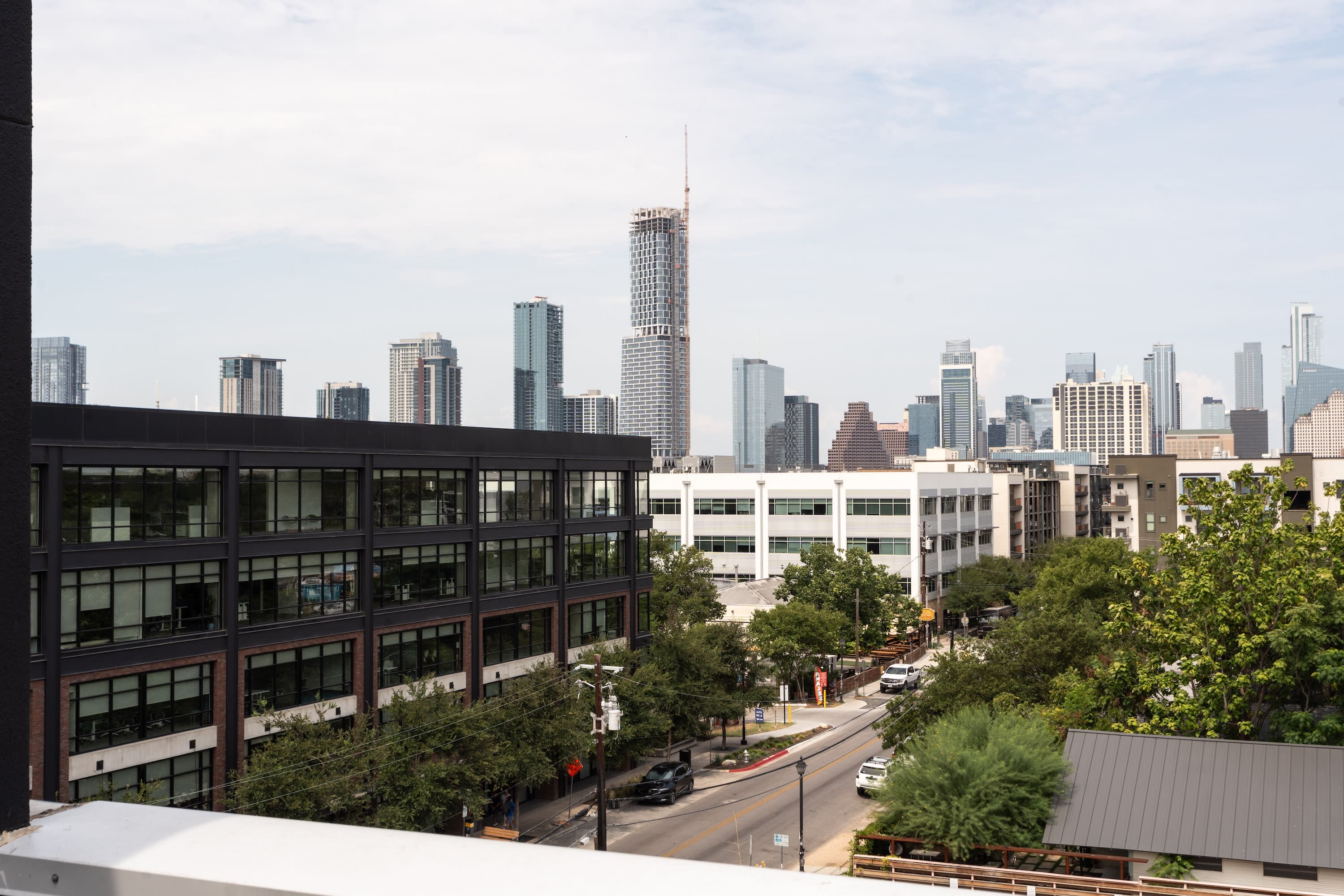 A modern dark-colored office building and a street with greenery occupy the foreground, while a dense city skyline with various skyscrapers stands under a pale sky in the background.