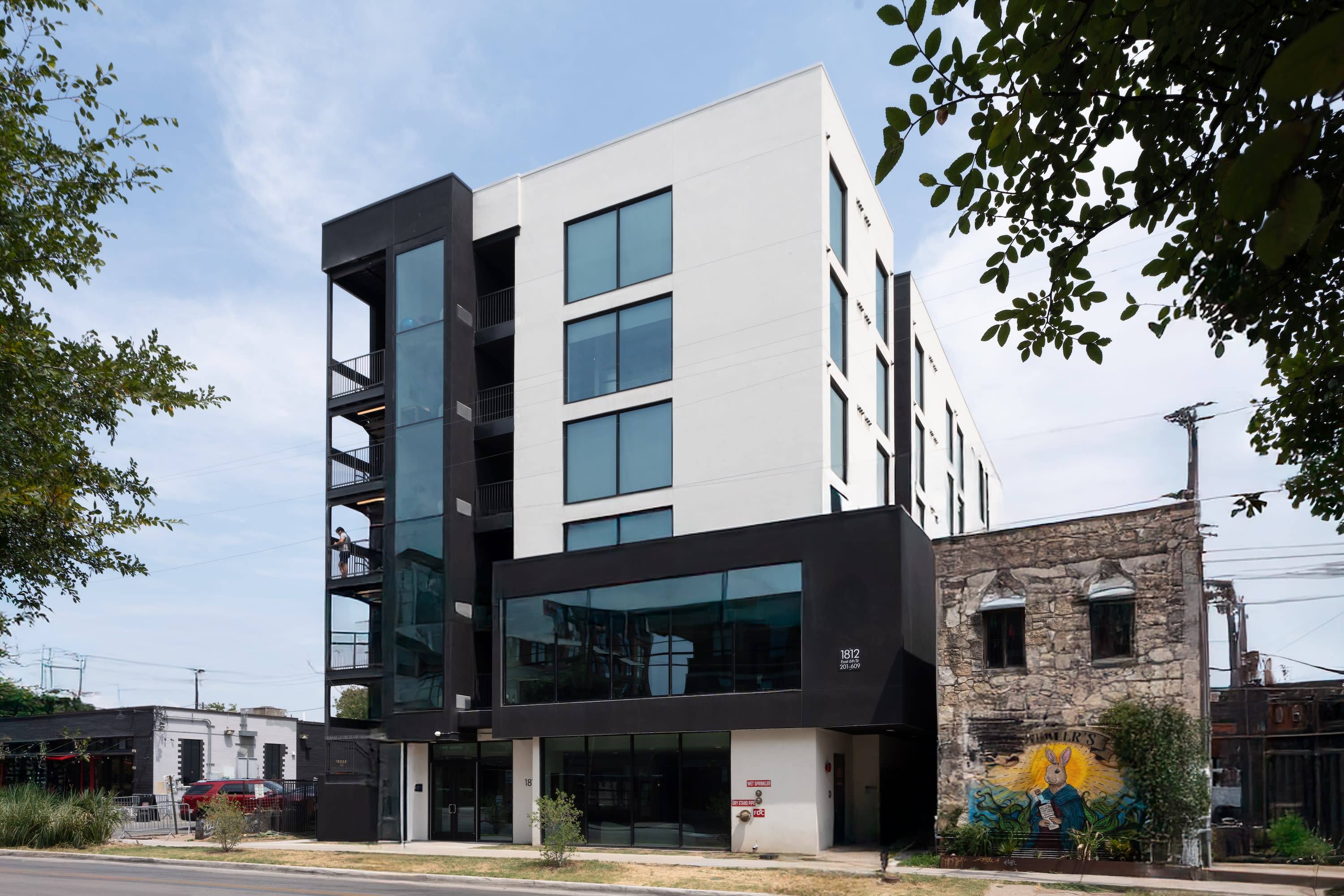 A modern black and white multi-story building stands next to an older stone structure with a colorful mural, set against a clear blue sky.