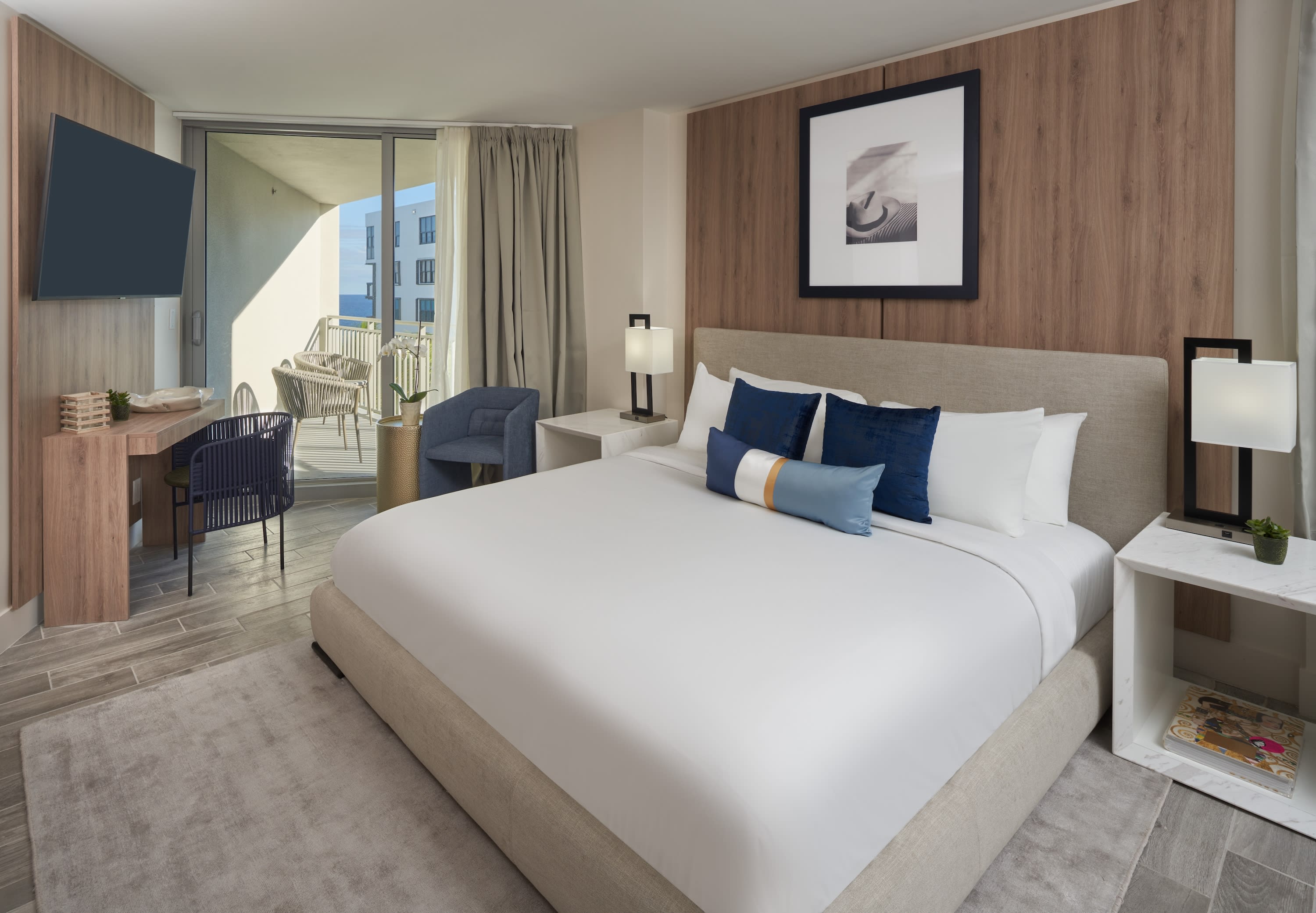 A large bed with white linens and blue pillows sits in the foreground of a modern hotel room, with a desk, seating area, and a balcony visible in the background.