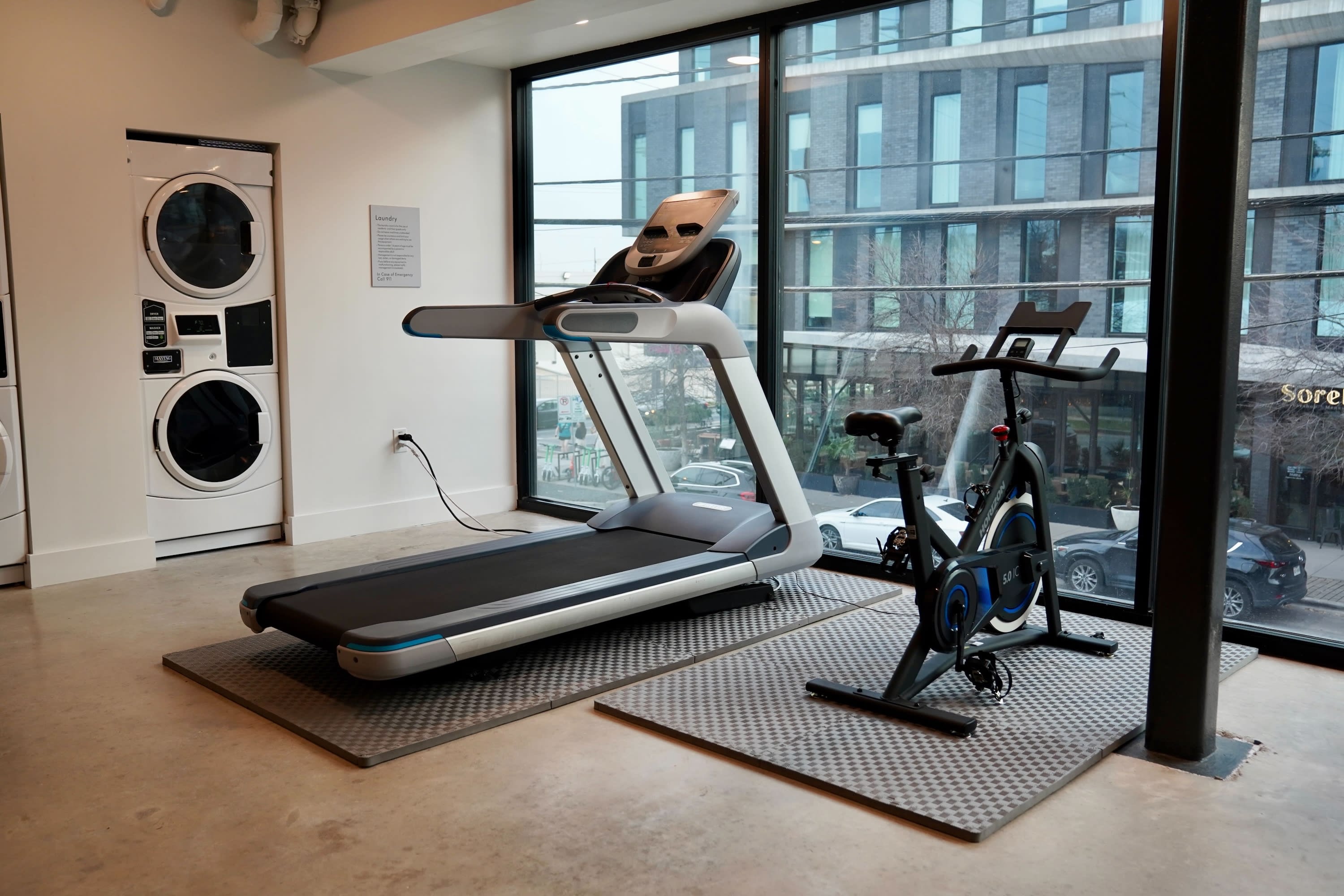 A treadmill and a stationary bike sit on rubber mats in a room with a laundry area and large windows overlooking a city building.