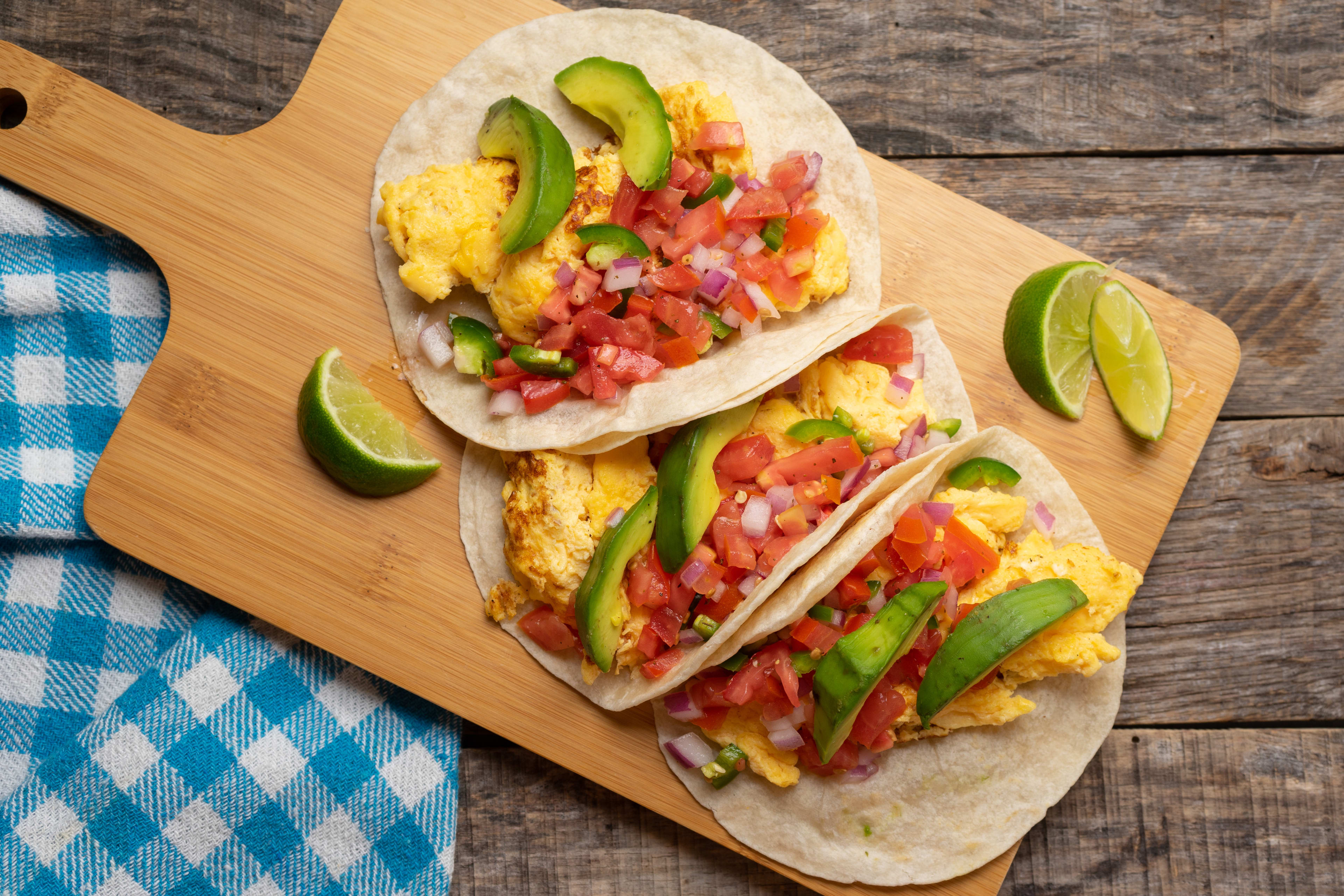 Three breakfast tacos filled with eggs, avocado, and pico de gallo sit on a wooden cutting board alongside lime wedges, resting on a rustic wooden table with a blue checkered napkin.
