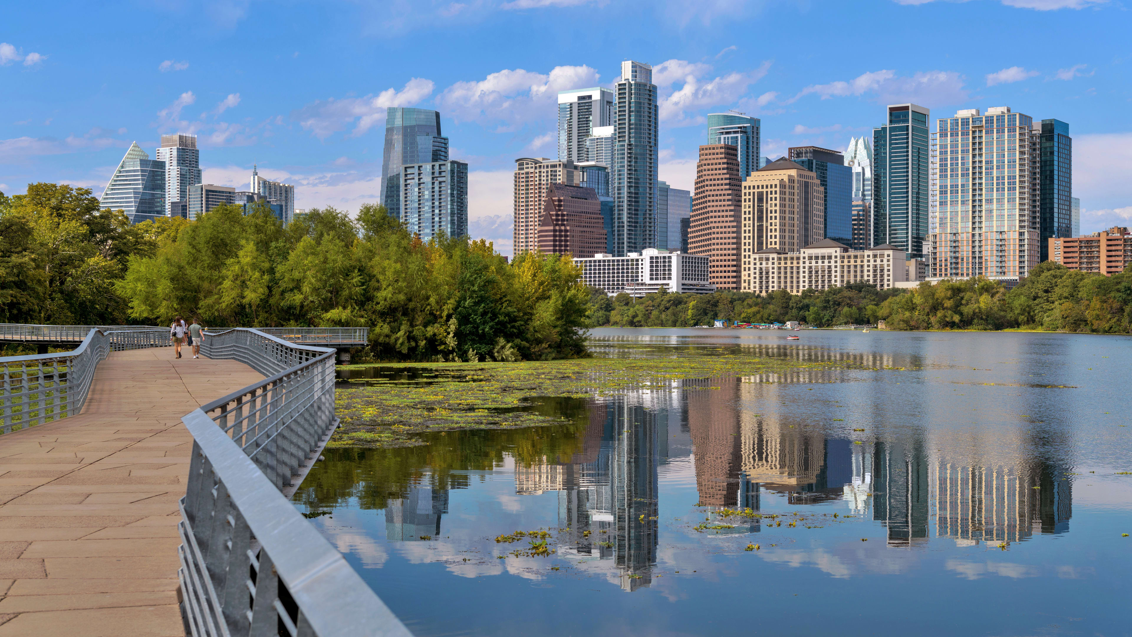 A concrete walkway with metal railings leads toward a lush green shoreline, with a city skyline reflecting in the calm water under a blue sky.