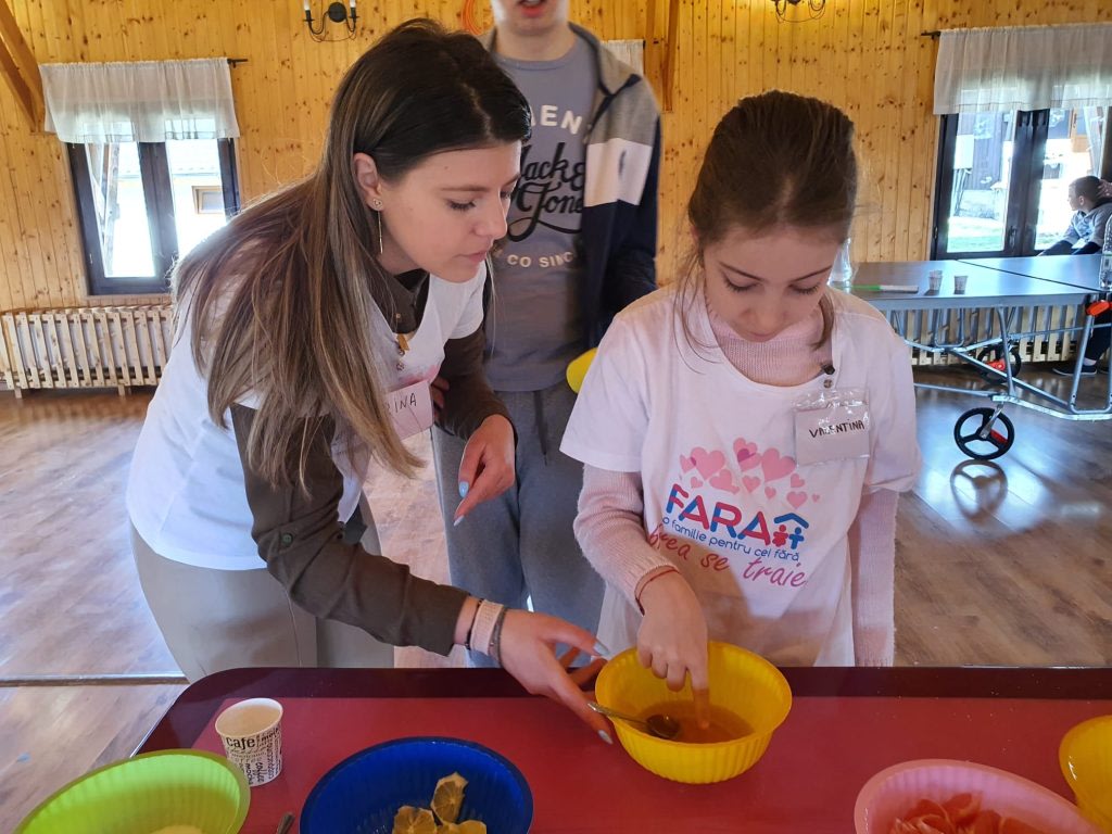 O voluntară ajută o fetiță la un atelier de bucătărie, într-o sală de lemn din tabără
