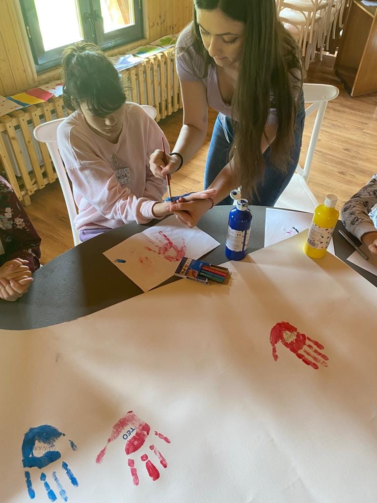 O voluntară pictează cu pensula palma unei fete, la un atelier; pe masă, foi cu amprente de palme roșii și albastre și sticluțe de tempera