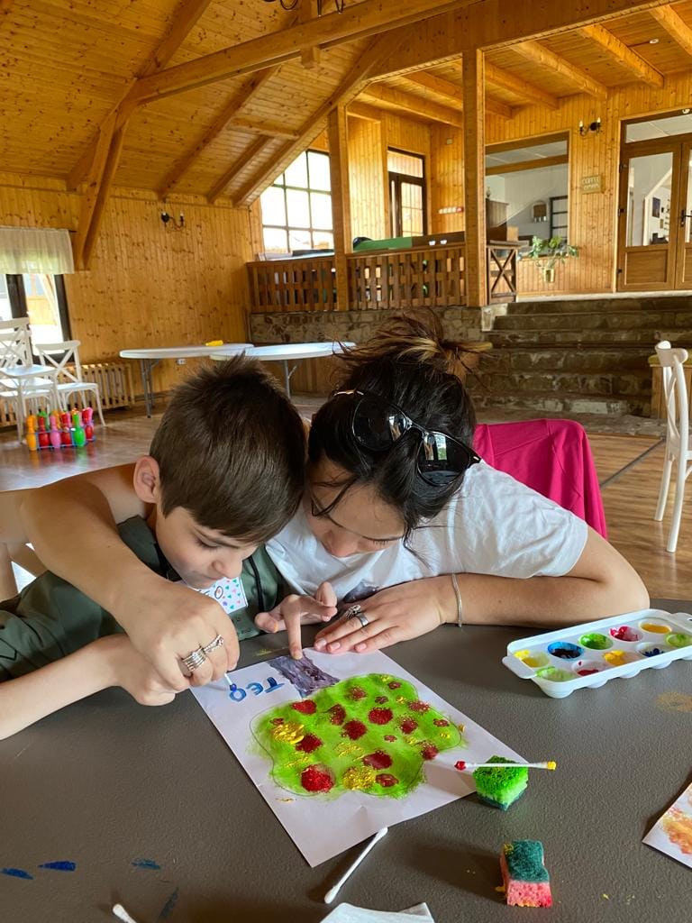 Un băiat și o voluntară, cu capetele apropiate, pictează împreună o foaie la masă