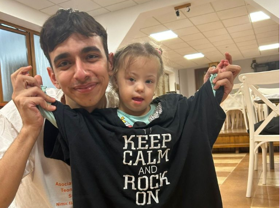 Un voluntar ține în fața unei fetițe un tricou negru cu „Keep calm and rock on”; amândoi zâmbesc