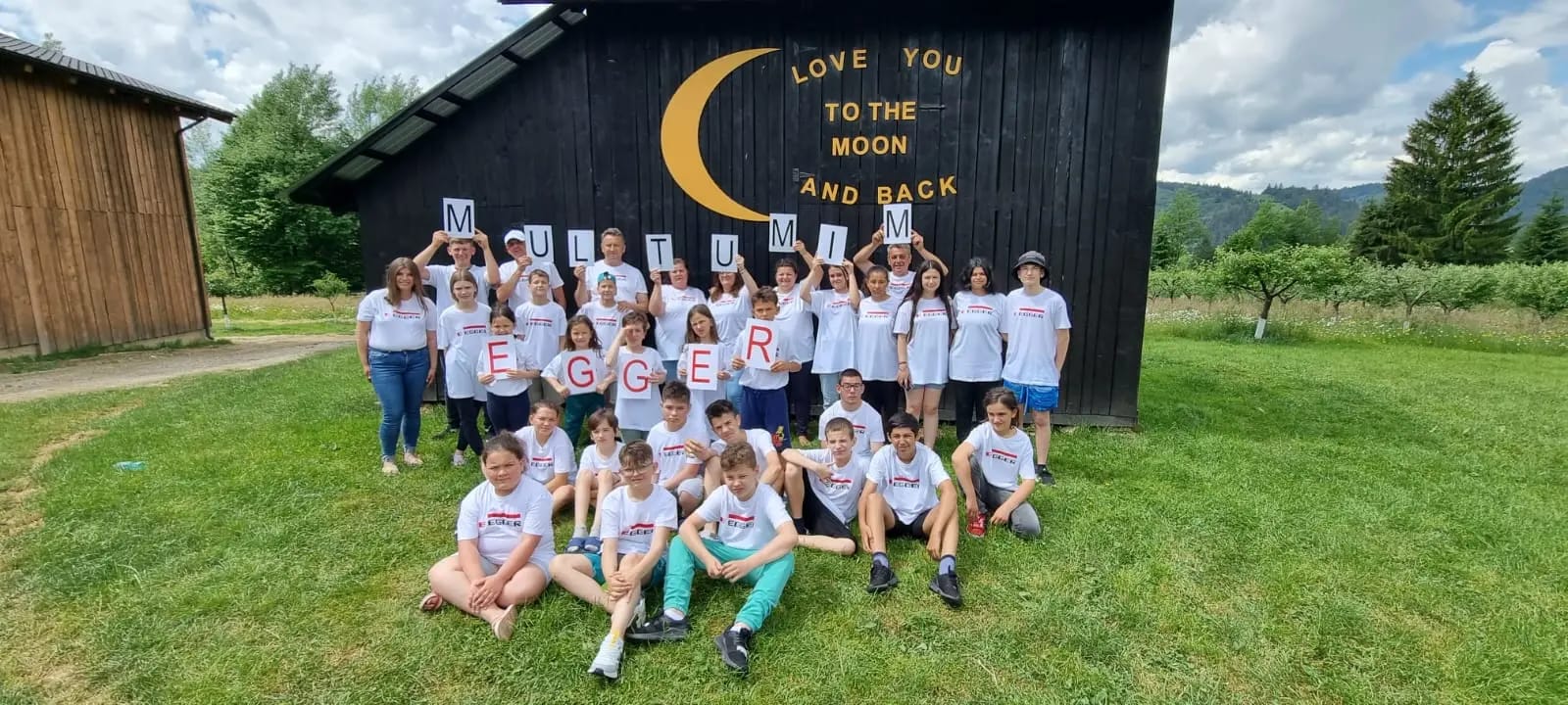 Copii și adulți în tricouri EGGER țin litere care formează „Mulțumim Egger”, în fața unui hambar de lemn negru cu o lună aurie și textul „Love you to the moon and back”