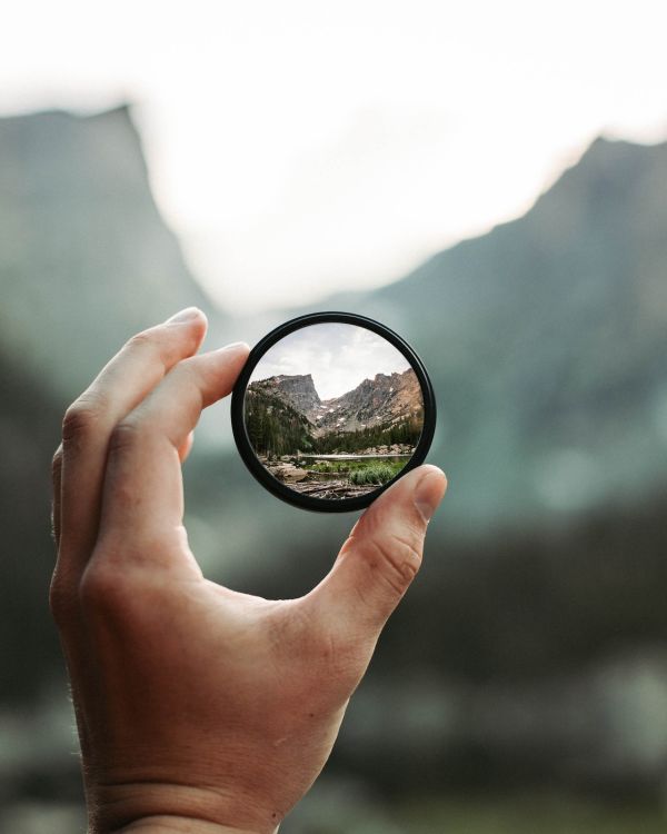 A view through a round lens being held at arms length. Visible through the lens is a crisp image of two rocky mountain peaks, pine trees, and a lake. A silhouette of the mountain peaks is visible but out of focus behind the lens.
