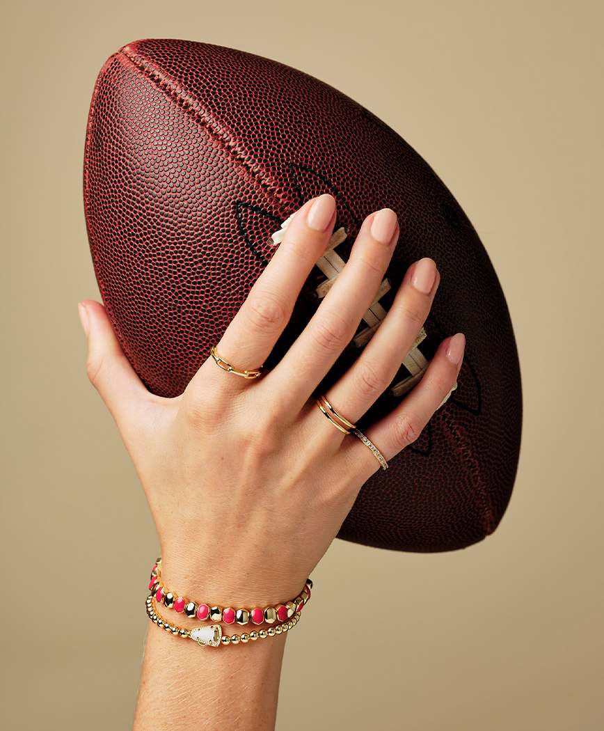 woman holding football wearing Kendra Scott daphne enamel gold stretch bracelet, along with other gameday bracelets and rings