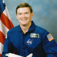 Astronaut Bruce Melnick in blue flight suit standing with arms crossed in front of space shuttle Atlantis