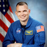Fotografía del astronauta Tony Antonelli con traje de vuelo azul delante del fla de EE.UU.