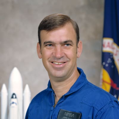 Headshot of astronaut John Fabian in blue flight suit in front of a space shuttle model and flag with official NASA seal