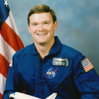 Astronaut Bruce Melnick in blue flight suit standing with arms crossed in front of space shuttle Atlantis