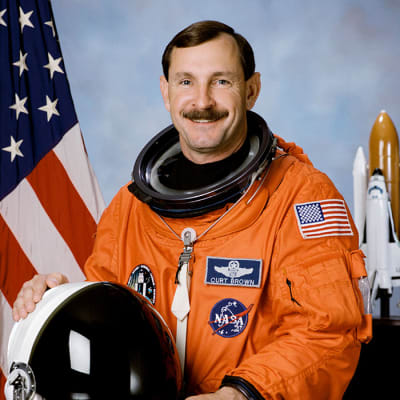 Headshot of astronaut Curt Brown in orange space suit holding his helmet in front of a U.S. flag and model of the space shuttle