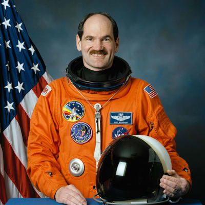 Headshot of astronaut Guy Gardner in orange flight suit holding helmet
