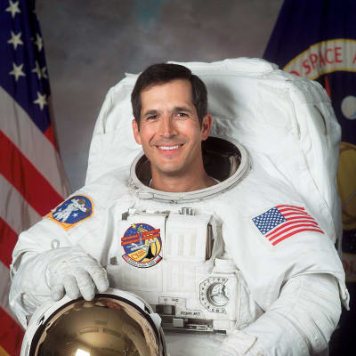 Headshot of astronaut Commander John Herrington in full space suit, holding his helmet in front of the U.S. and NASA flags