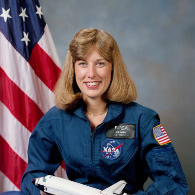 Headshot of astronaut Jan Davis in blue flight suit holding a model space shuttle in front of the U.S. flag