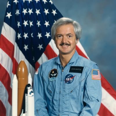 Astronaut John Bartoe in blue flight suit in front of the American flag and next to a model of the space shuttle