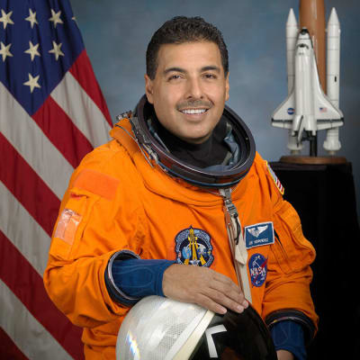 Headshot of astronaut José Hernandez in orange space suit, holding his helmet and standing in front of the U.S. flag and a model of the space shuttle
