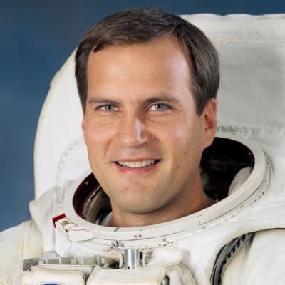 Astronaut Richard Hieb in full space suit holding his helmet in front of U.S. flag