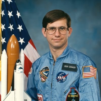 Photo of NASA astronaut Robert Cenker next to the United States flag and a model of the space shuttle