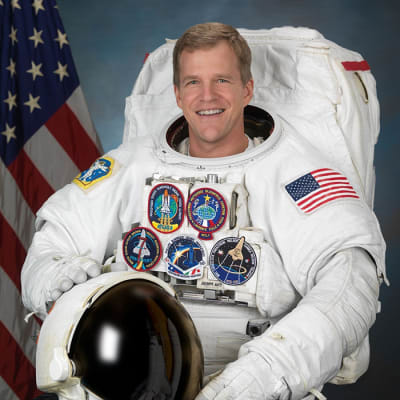 Astronaut Scott Parazynski in full space suit holding his helmet in front of the American flag