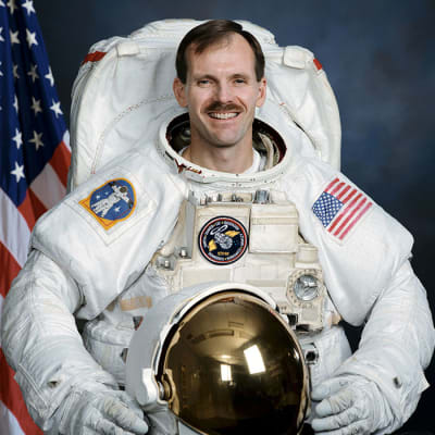 Astronaut Steven Smith in full space suit holding his helmet in front of the U.S. flag