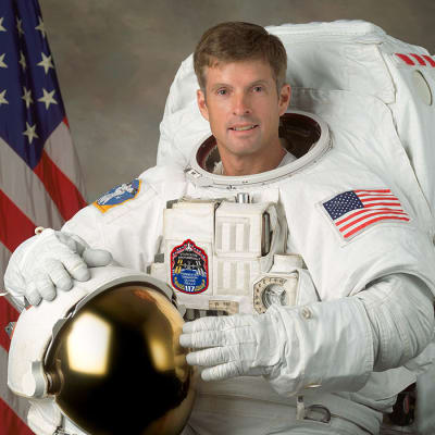 Headshot of astronaut Steve Swanson in full space suit holding his helmet in front of the U.S. flag