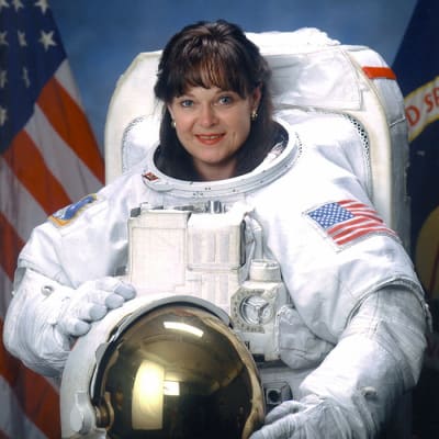 Headshot of astronaut Tammy Jernigan in full space suit holding her helmet in front of the NASA flag and U.S. flag